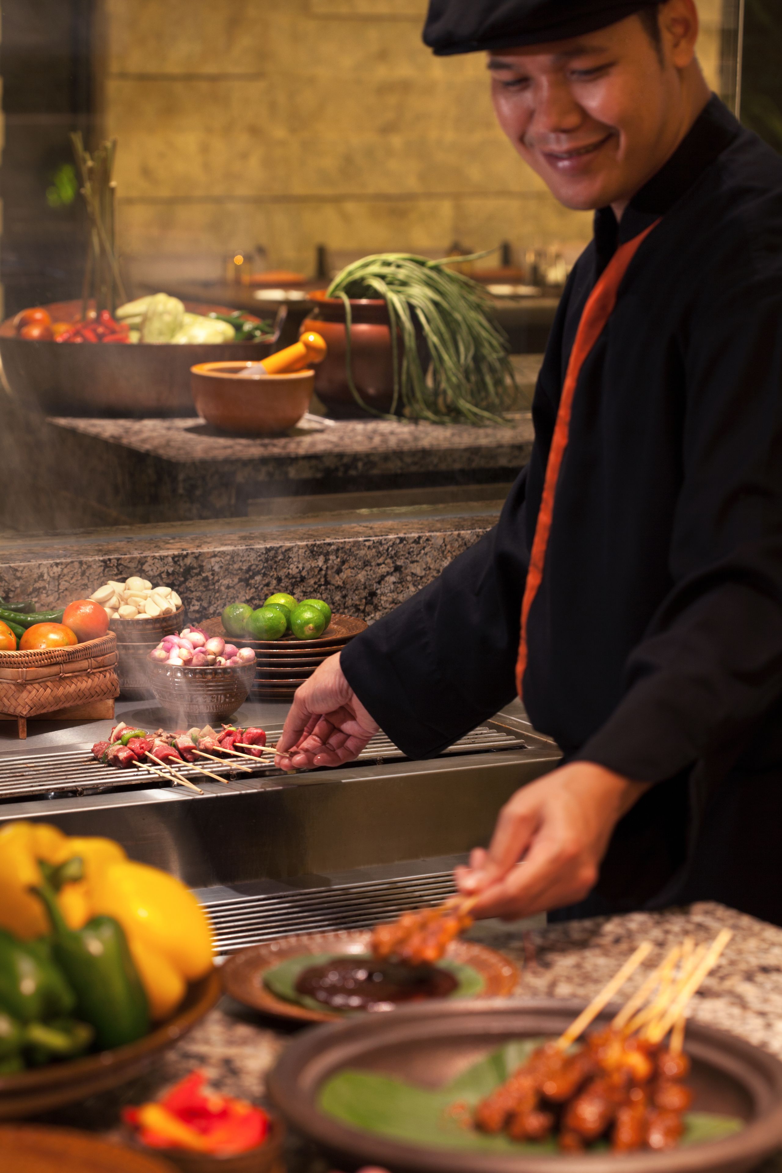 Un chef preparando brochetas con verduras y carne, rodeado de diversos ingredientes y utensilios de cocina.