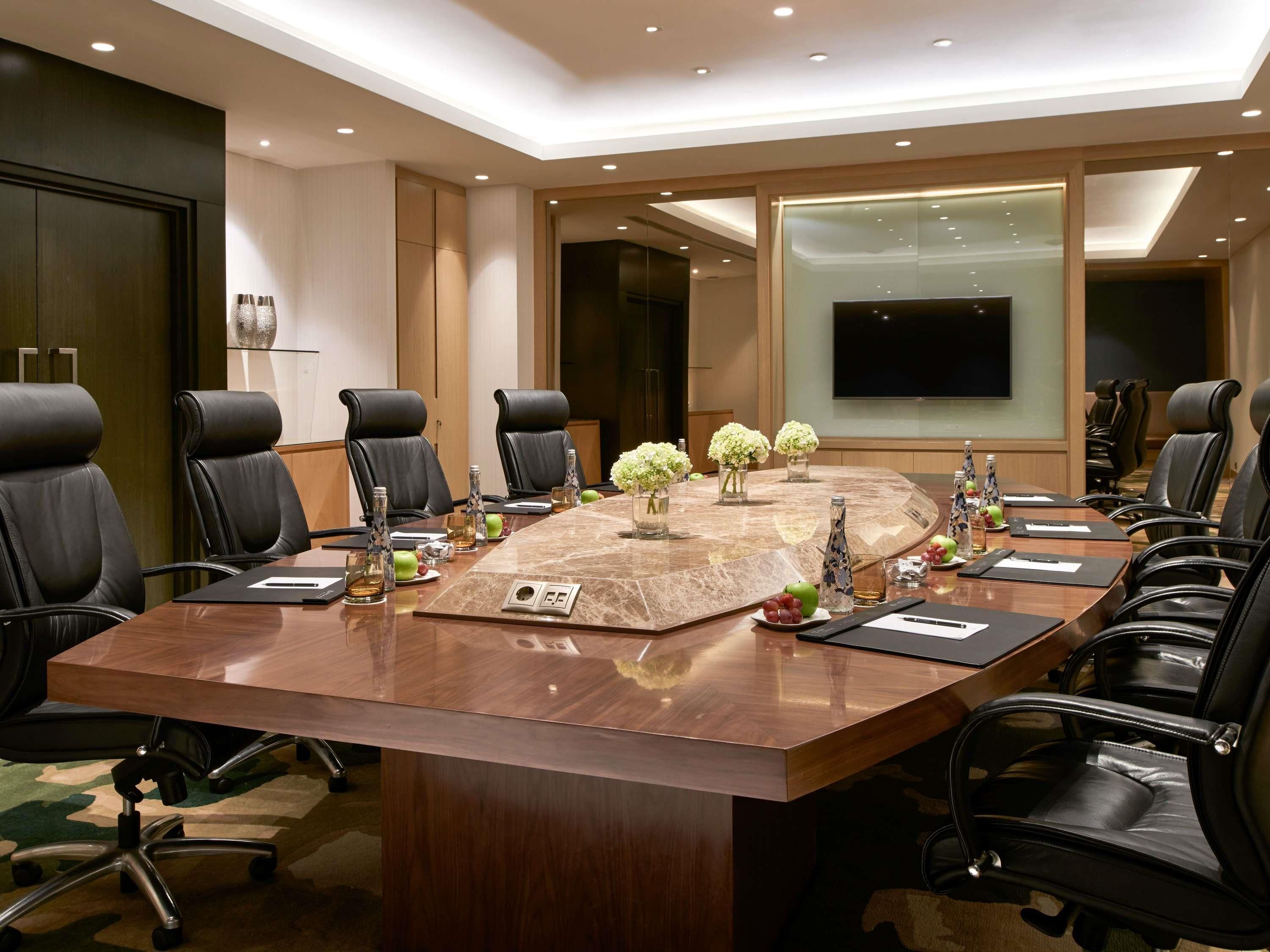 Una sala de conferencias con una gran mesa ovalada, sillas de cuero negro, un televisor de pantalla plana y jarrones decorativos.