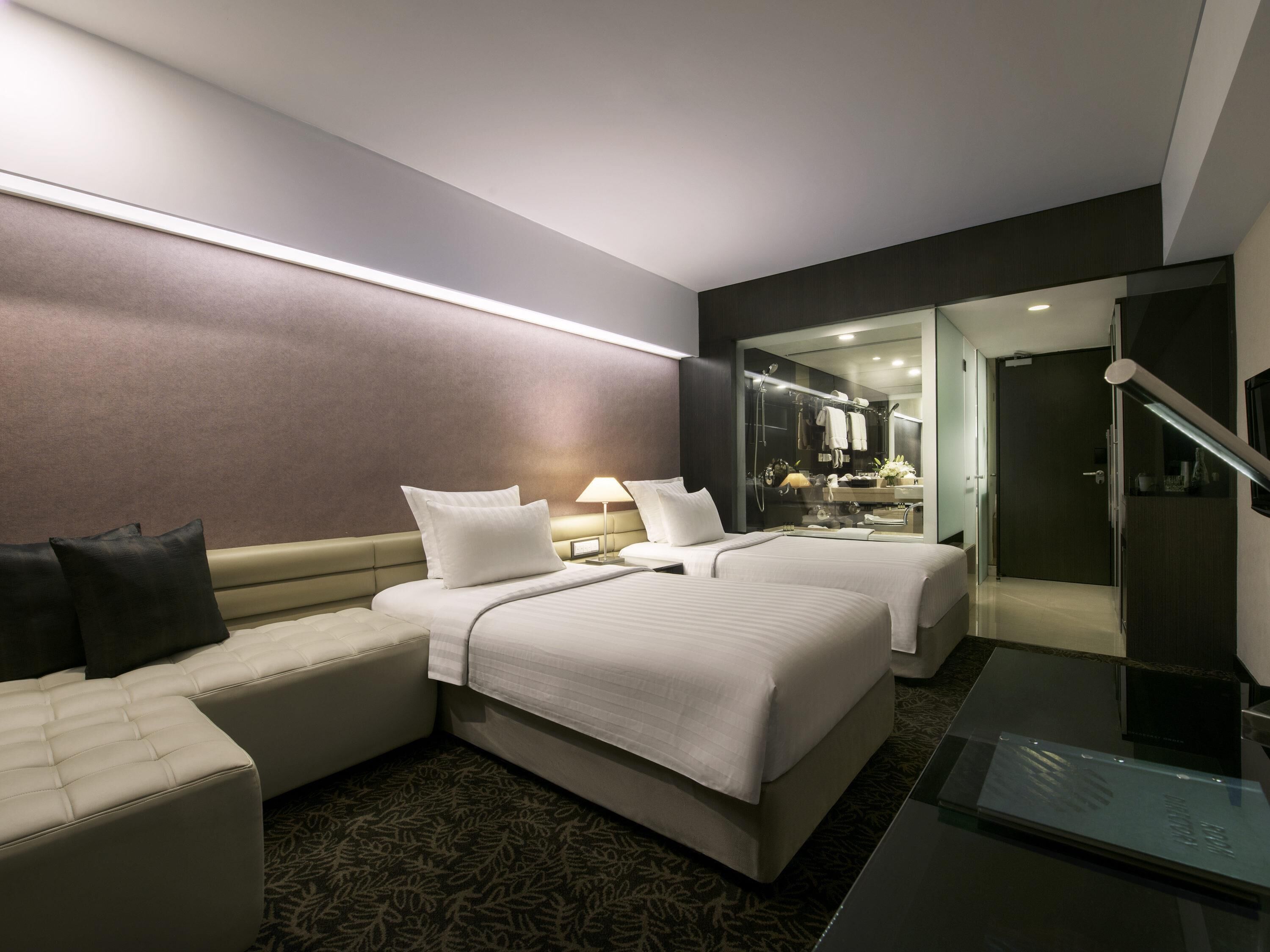 A hotel room with two beds, a sofa, a glass shower enclosure, and a separate bathroom visible through a glass door.