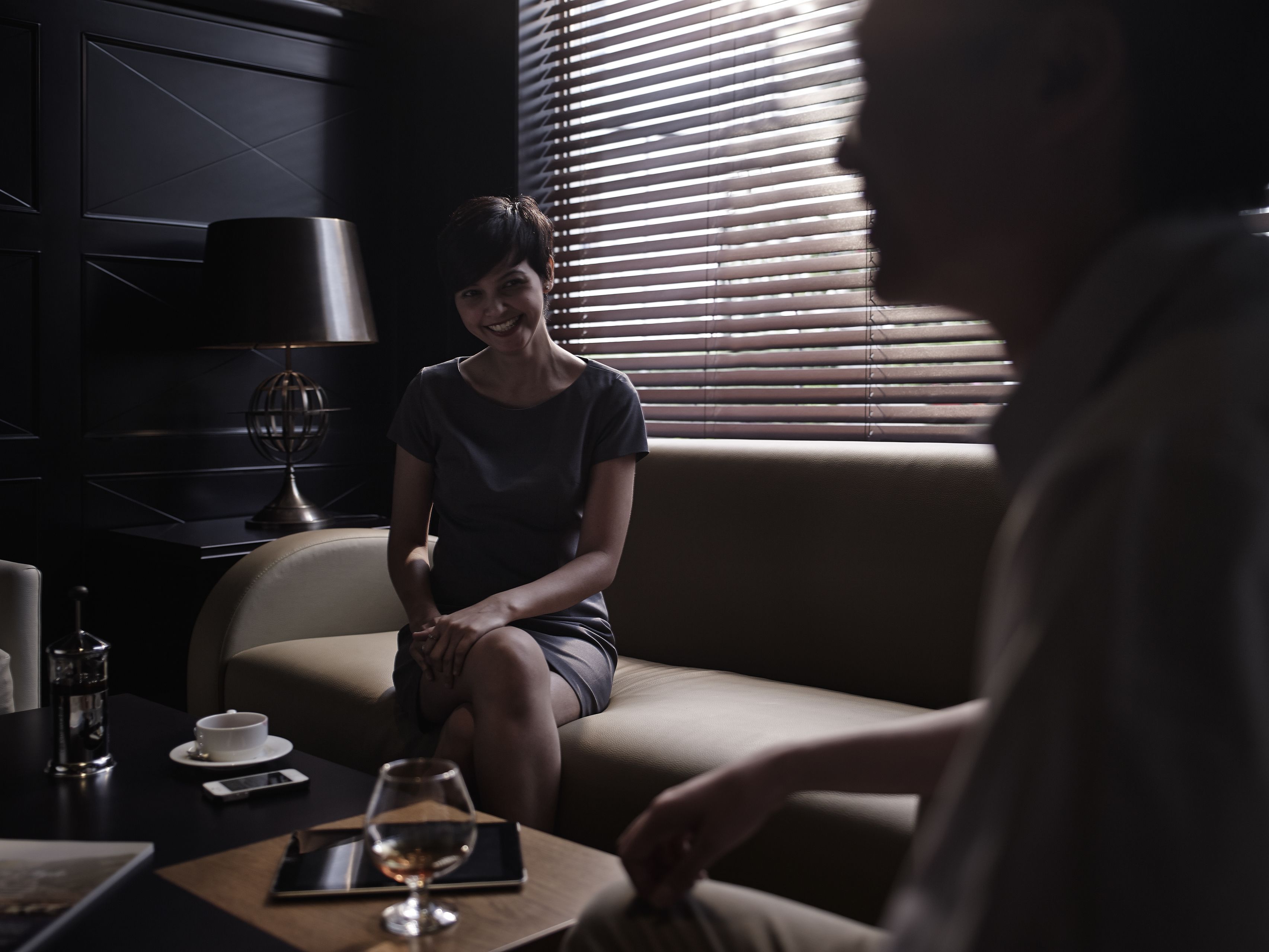 A dimly lit room with a person sitting on a couch, a table with a glass of wine, and a lamp.