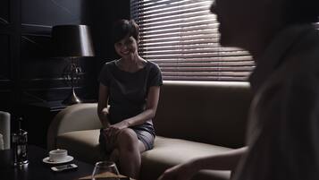 A dimly lit room with a person sitting on a couch, a table with a glass of wine, and a lamp.