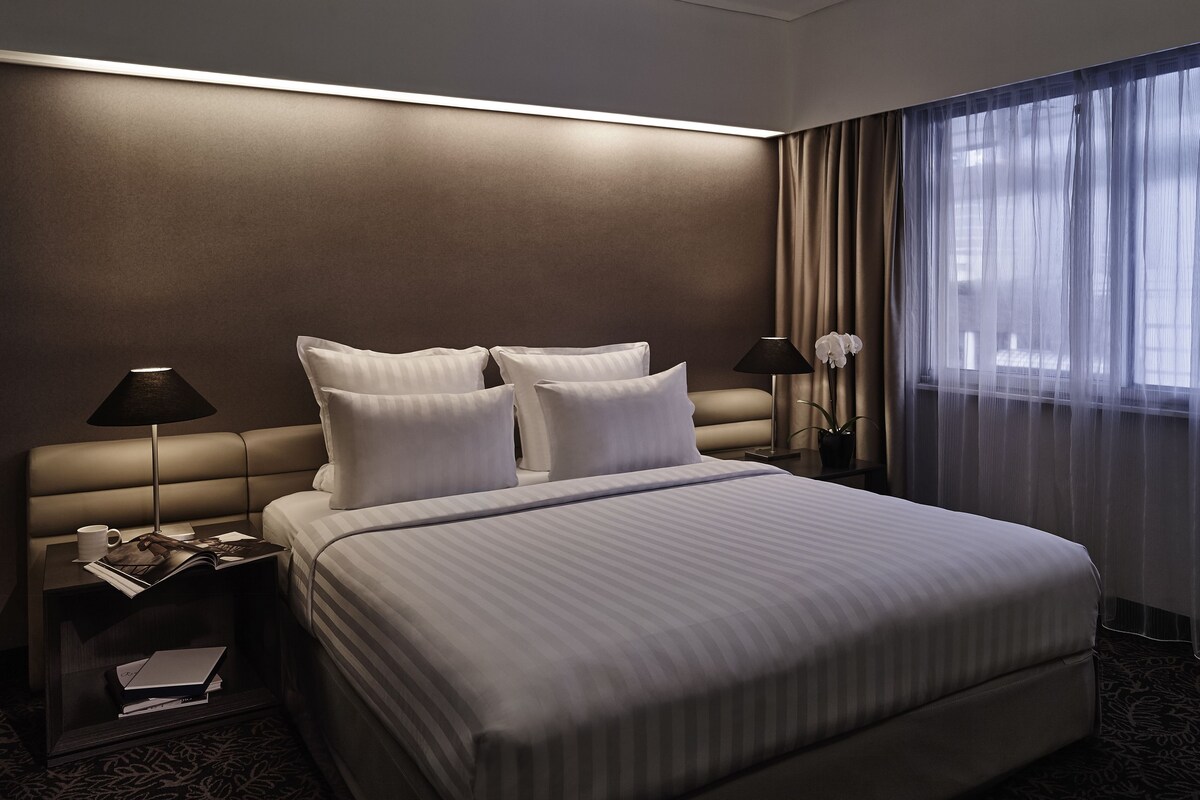 Habitación de hotel con una cama grande, dos mesitas de noche, una lámpara, una planta en maceta y una ventana con cortinas.