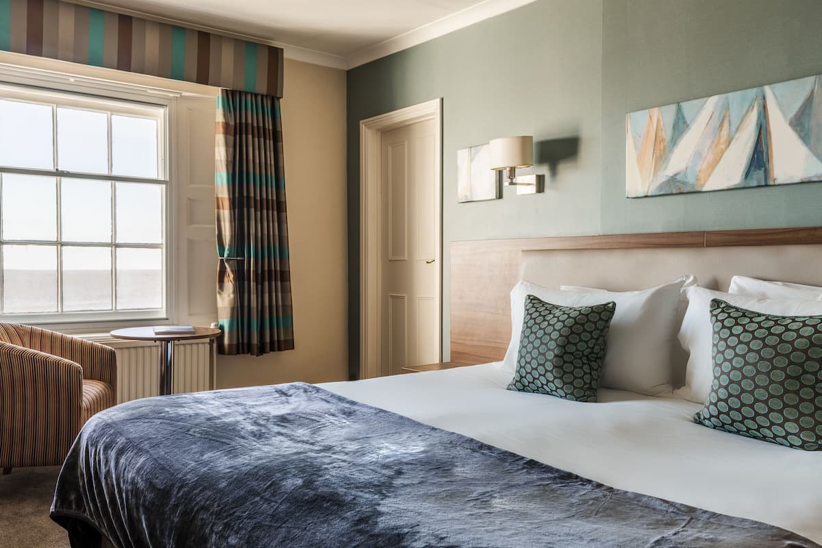 A hotel room with a large bed, a chair, a window with curtains, a painting on the wall, and a lamp.