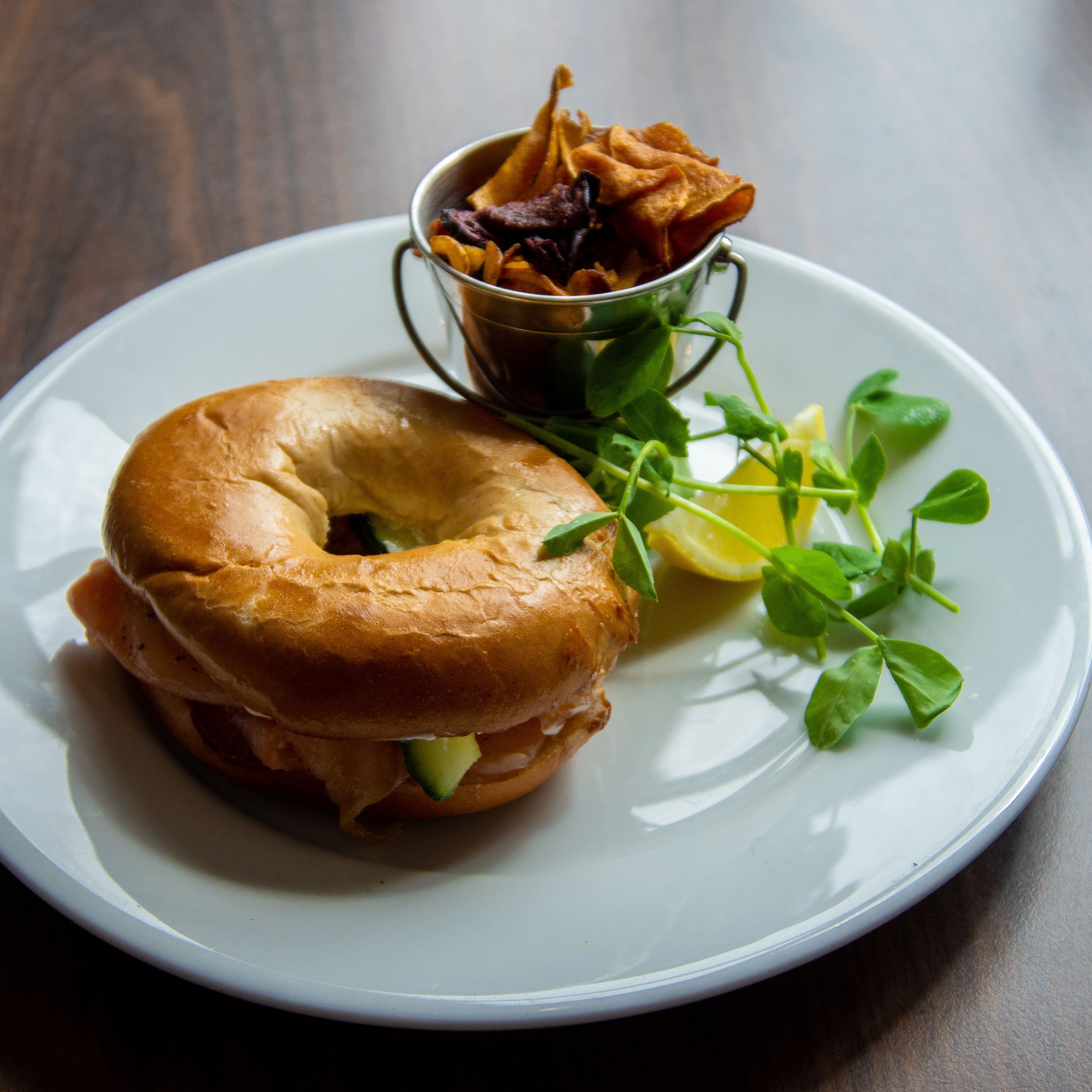 A plate with a bagel sandwich, a small metal cup with fries, and some greens.