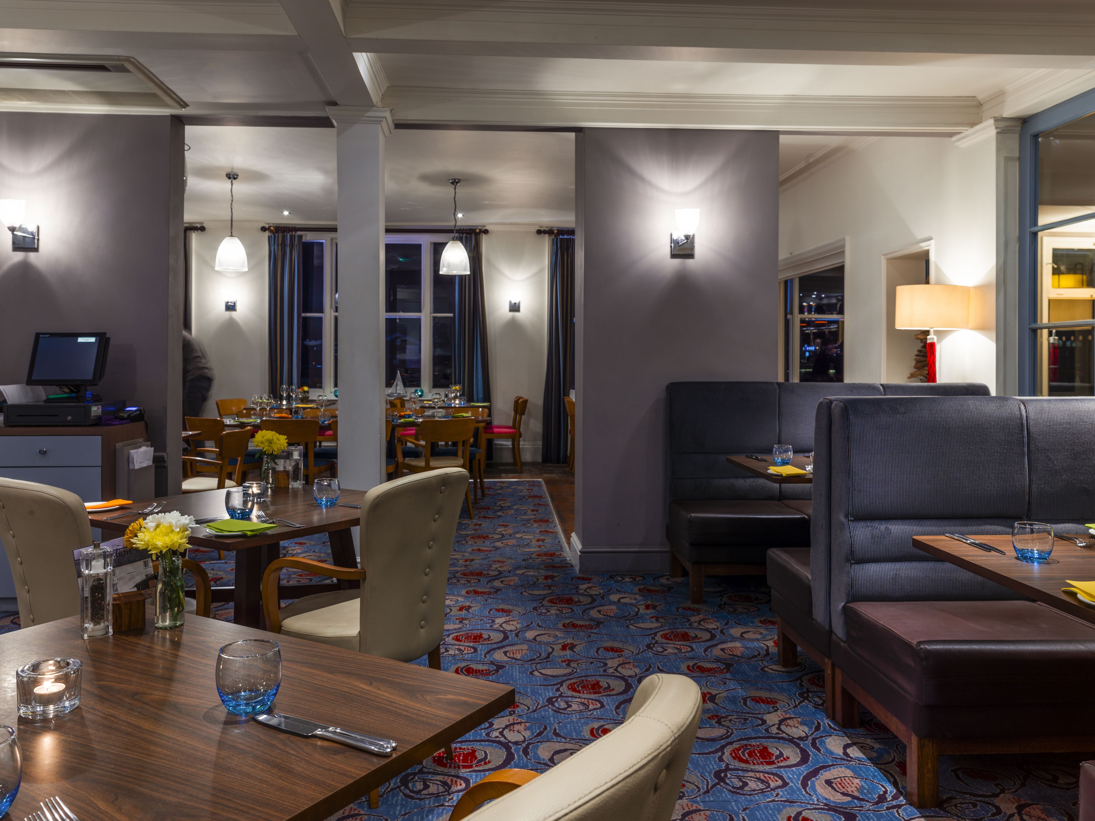 A hotel dining area with a long table, chairs, and a booth. There is a computer on a desk, and a window with curtains.
