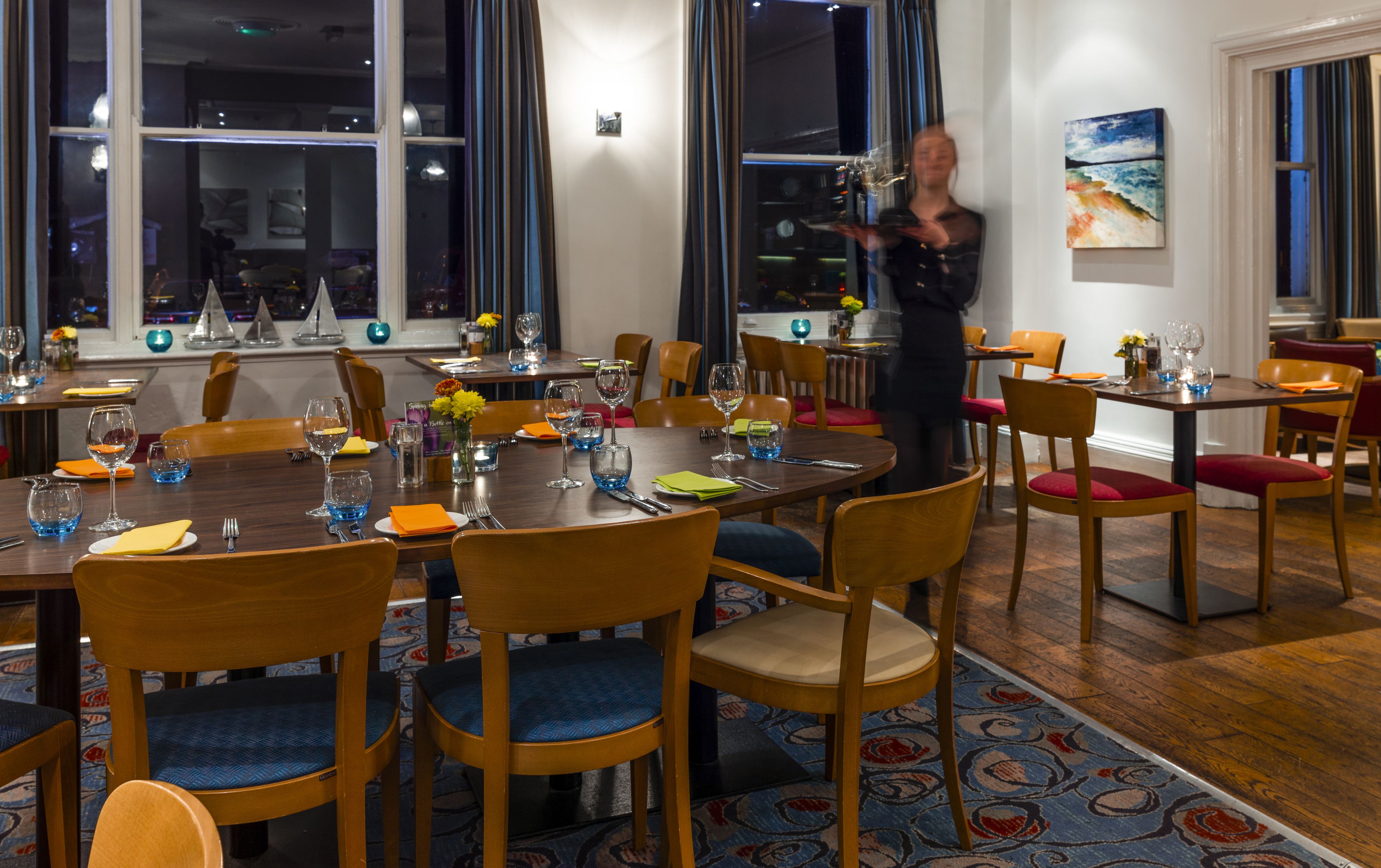 A dining area with wooden tables and chairs, set with plates, glasses, and cutlery.