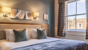 A hotel room with a large bed, two wall-mounted lamps, a painting, and a window with curtains.
