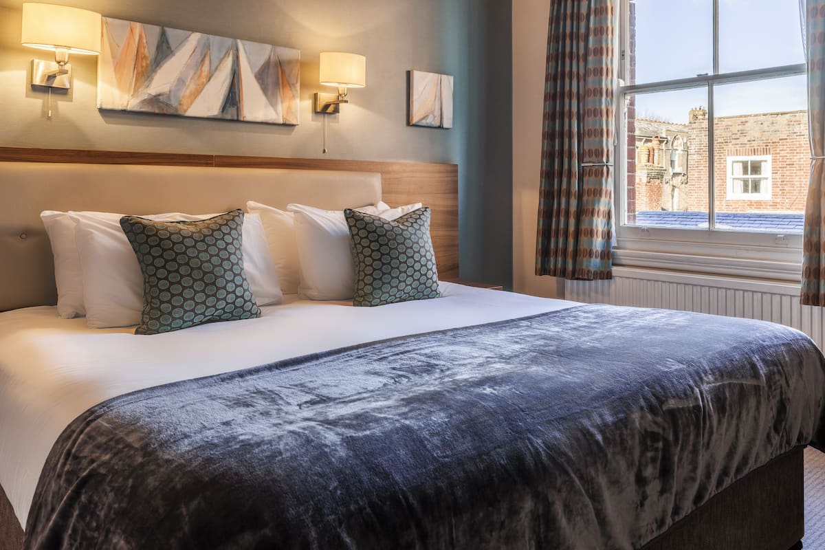 A hotel room with a large bed, two wall-mounted lamps, a painting, and a window with curtains.