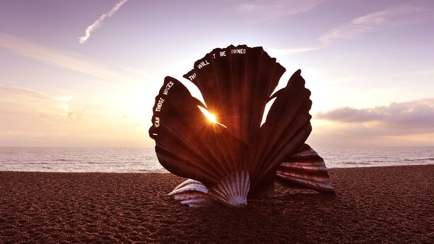 A large shell sculpture on a beach with the sun setting in the background.