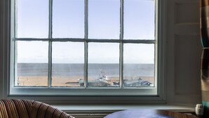 A room with a large window overlooking a beach and a ship. There is a chair and a table with a book on it.