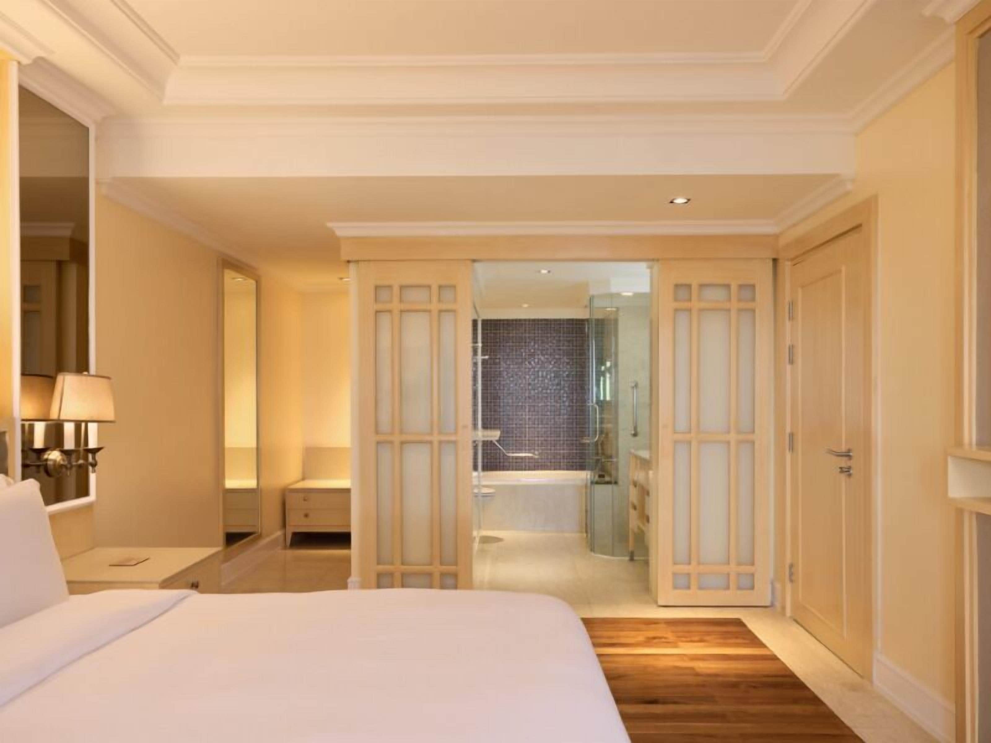 A hotel room with a large bed, a bedside table with a lamp, a wardrobe, and a bathroom visible through an open door.