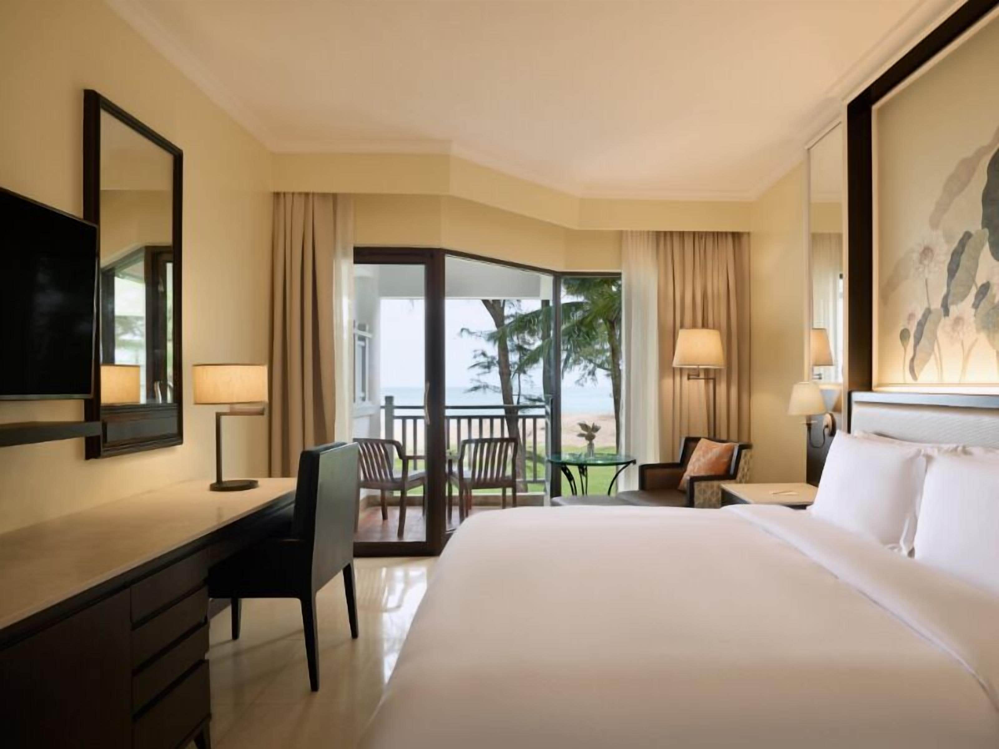 A hotel room with a large bed, a desk, a chair, a TV, and a balcony with a view of palm trees.