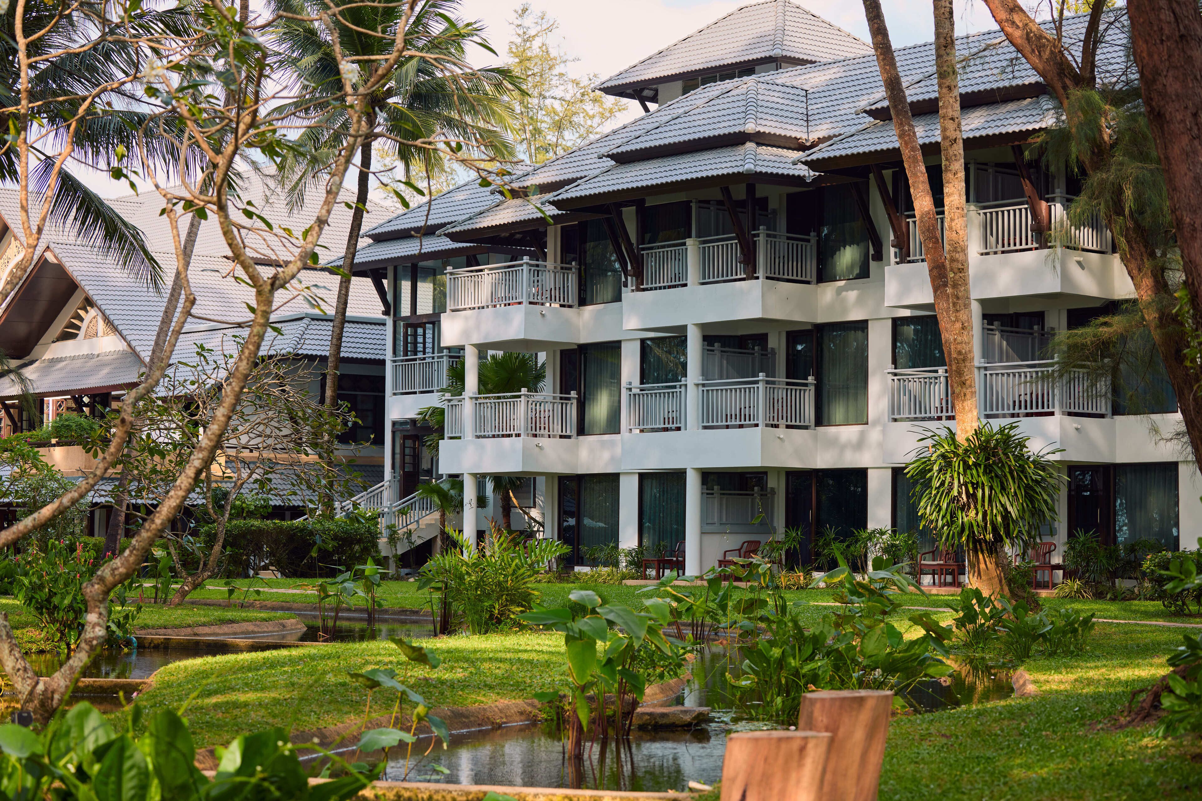 Un hotel de varios pisos con balcones, un jardín con un estanque y vegetación tropical.