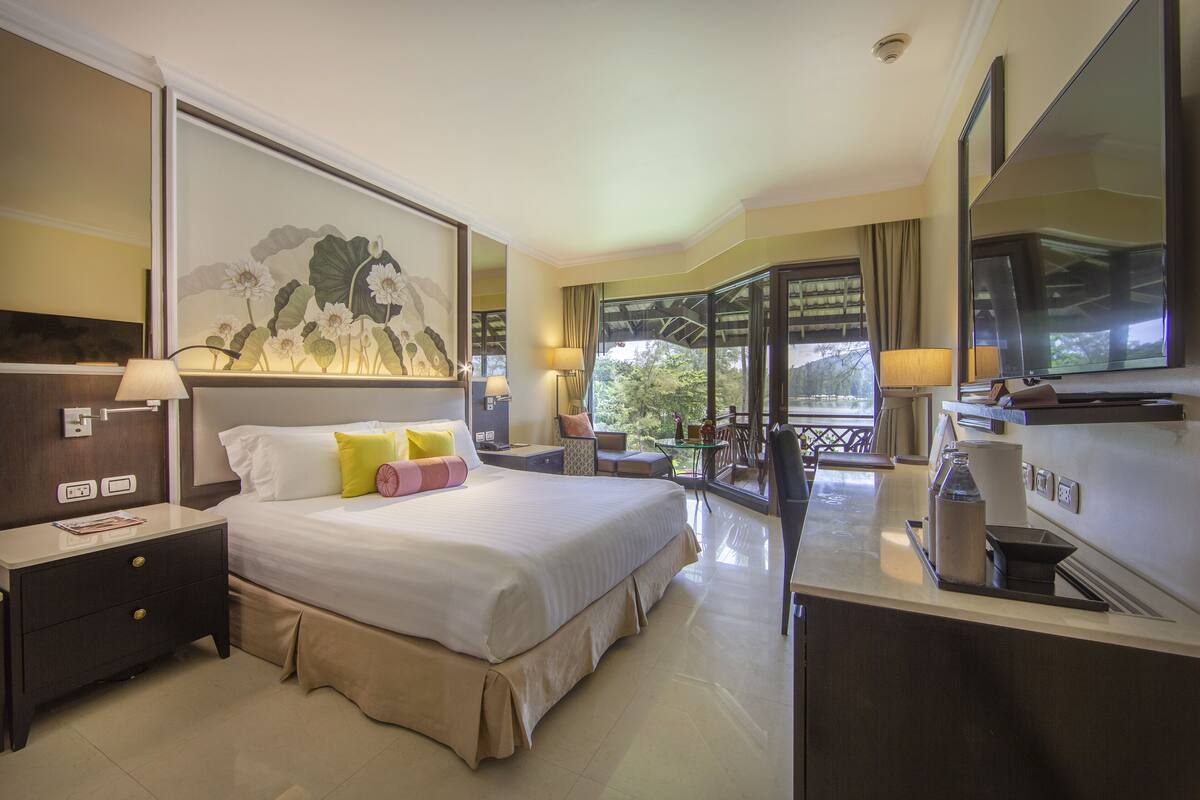 A hotel room with a large bed, a desk, a chair, a balcony with a view, and a painting on the wall.