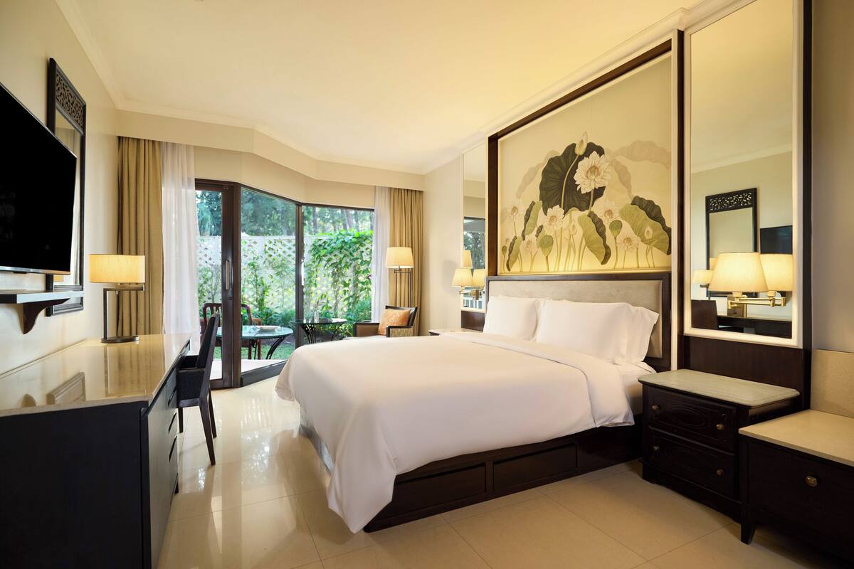 A hotel room with a large bed, a desk with a TV, a dining area with a table and chairs, and a balcony with a view of greenery.