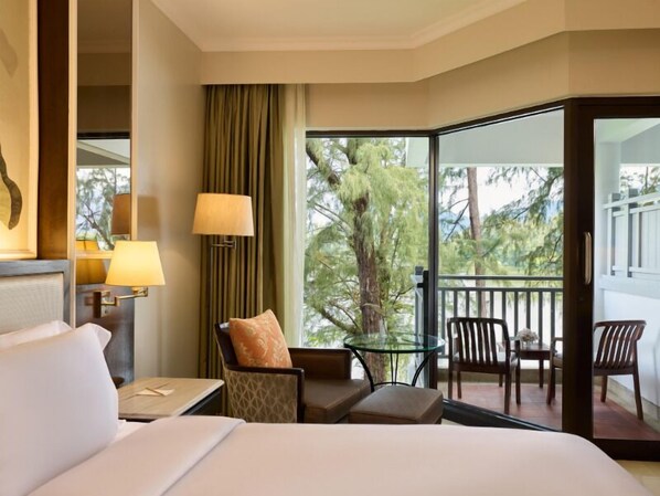 A hotel room with a bed, a desk, a chair, a glass table, and a balcony with a view of trees. - Dusit Thani Laguna Phuket (Choeng Thale)