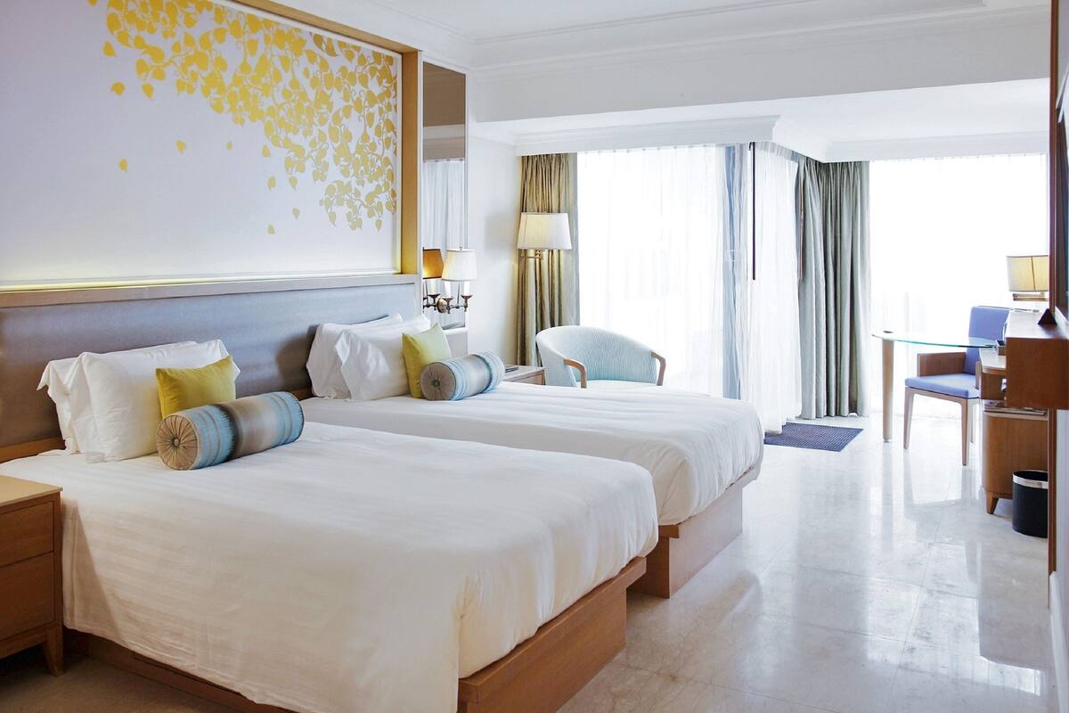 A hotel room with two beds, a desk, a chair, and a large window with curtains.