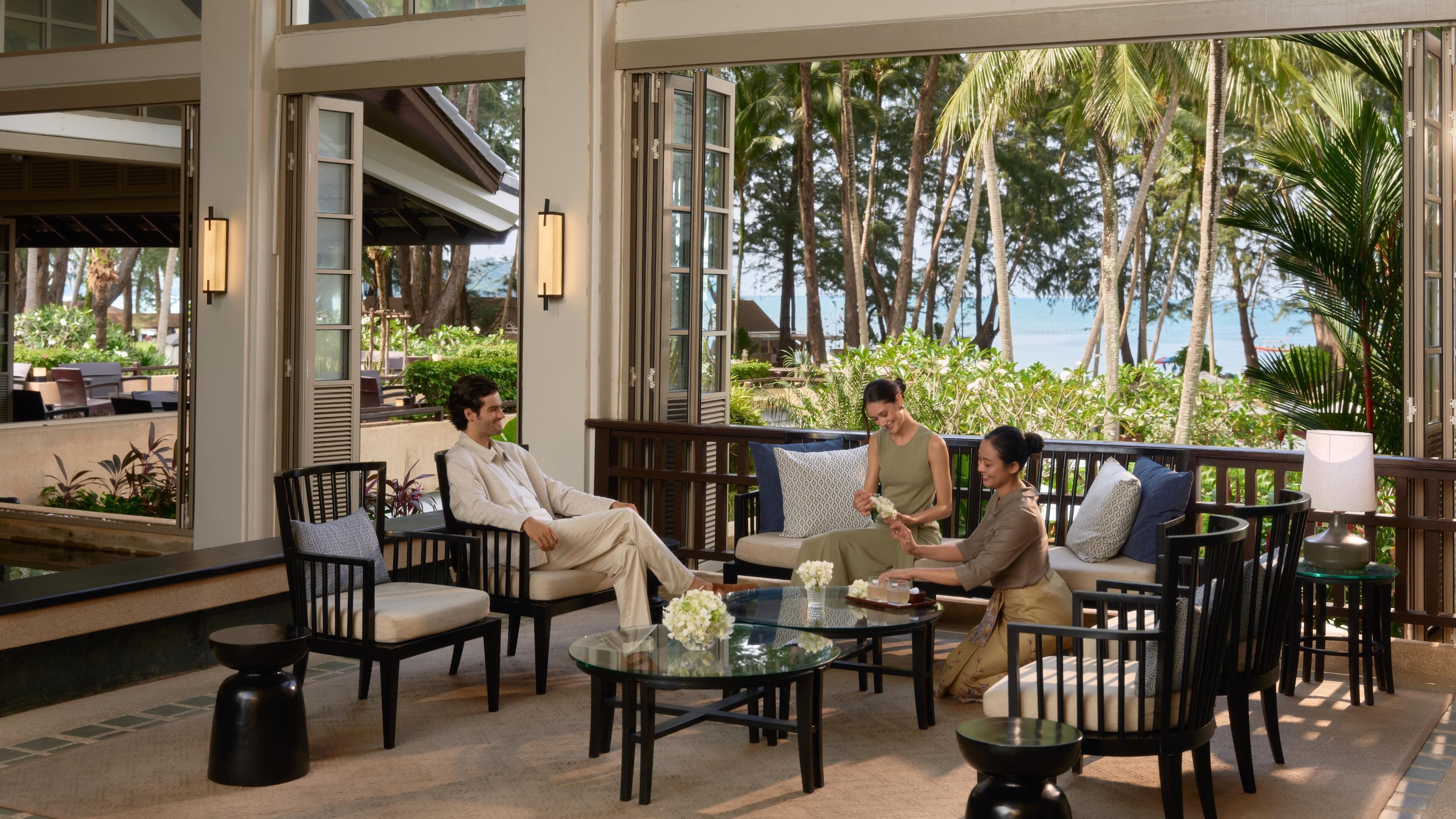 Un área de lounge al aire libre moderna con mesas de vidrio, asientos cómodos y vistas a palmeras y al océano.