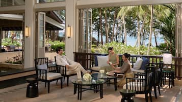 A modern outdoor lounge area with glass tables, comfortable seating, and a view of palm trees and the ocean.