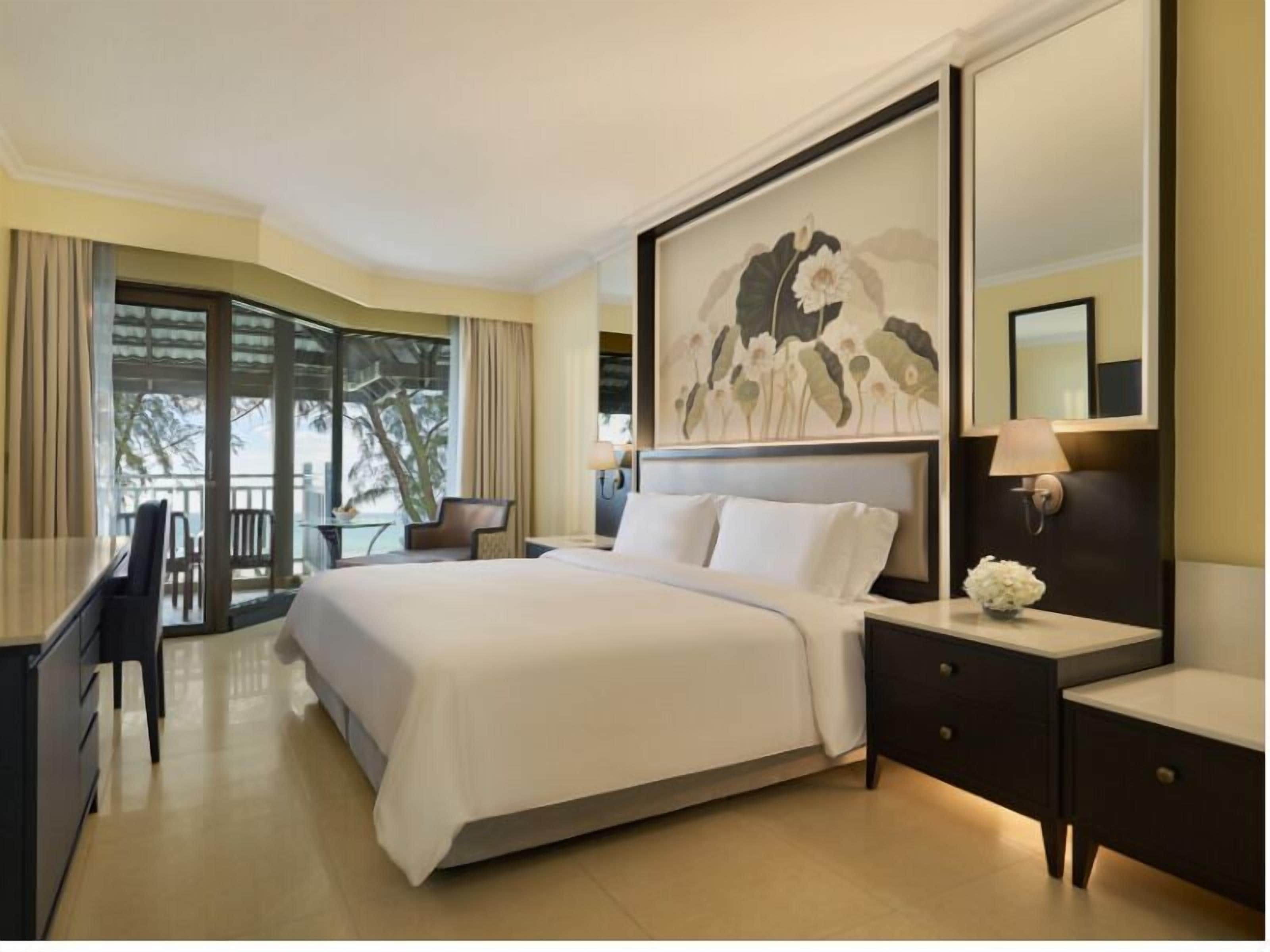 A hotel room with a large bed, a desk, a chair, a nightstand, and a window with a view of palm trees.