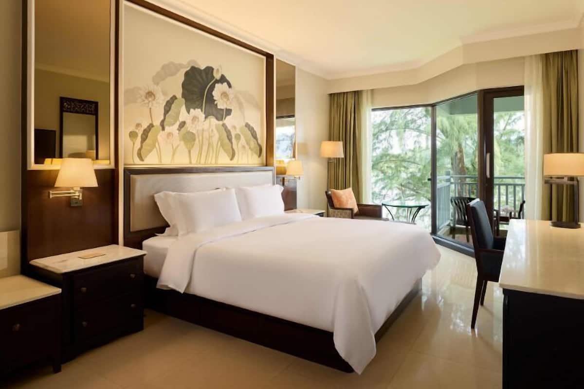 A hotel room with a large bed, a desk, and a view of greenery through the window.