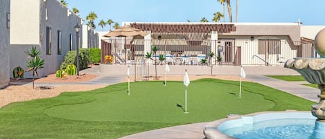 A golf course with a putting green, a fountain, and a building with a covered seating area.
