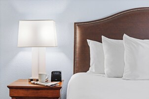 Ein ordentlich bezogenes Bett mit weißen Kissen und einem Holz-Kopfstück, ein Nachttisch mit Lampe, einer digitalen Uhr und einer Tasse.