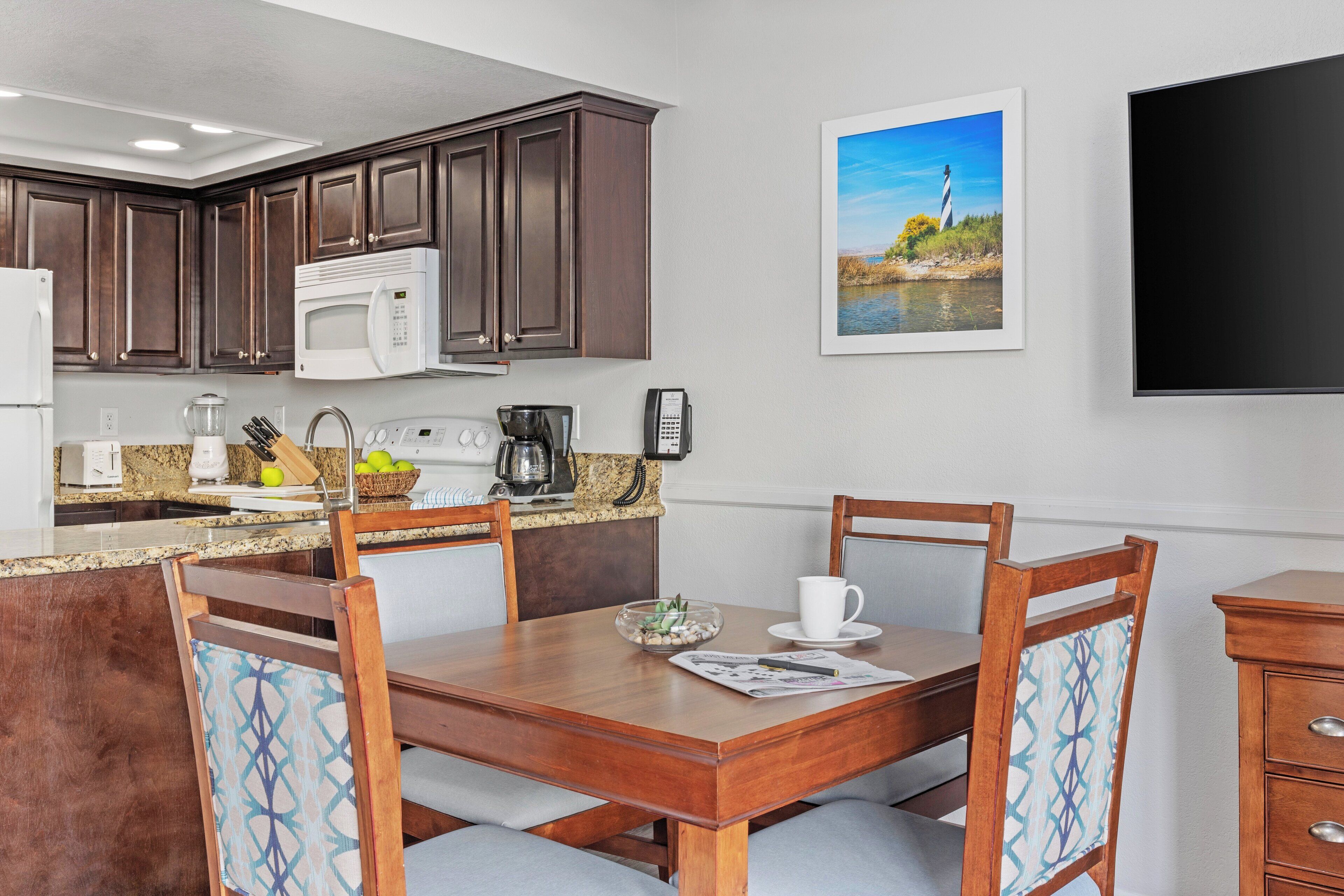 Una cocina moderna con armarios de madera oscura, un microondas blanco y una encimera de granito. Hay una mesa de comedor con cuatro sillas, una cafetera y un cuenco de fruta. En la pared, se observa un cuadro enmarcado de un faro, y un televisor de pantalla plana está montado sobre la encimera.