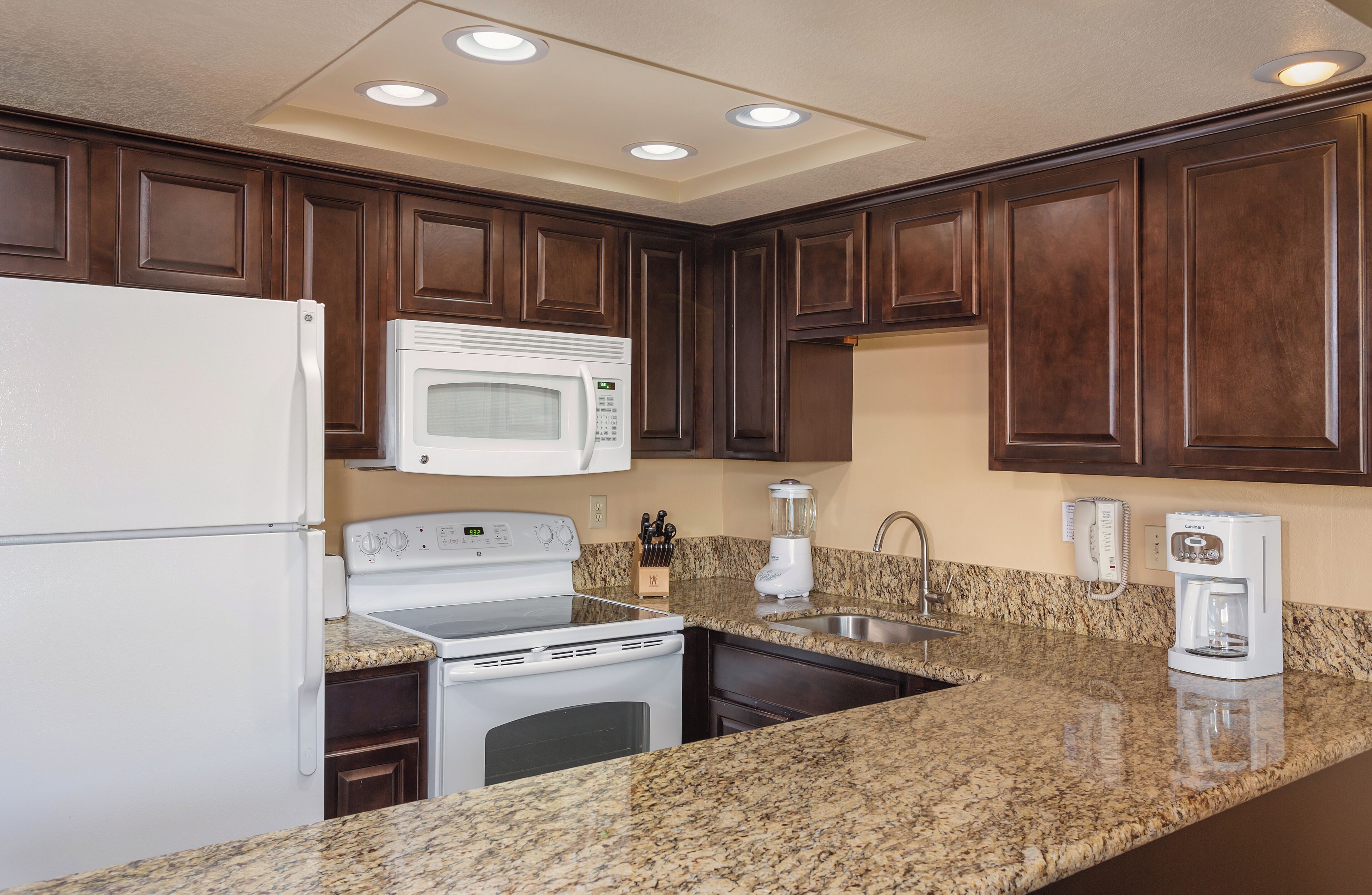 A kitchen with dark wooden cabinets, a white refrigerator, a microwave, a stove, a sink, and a coffee maker.