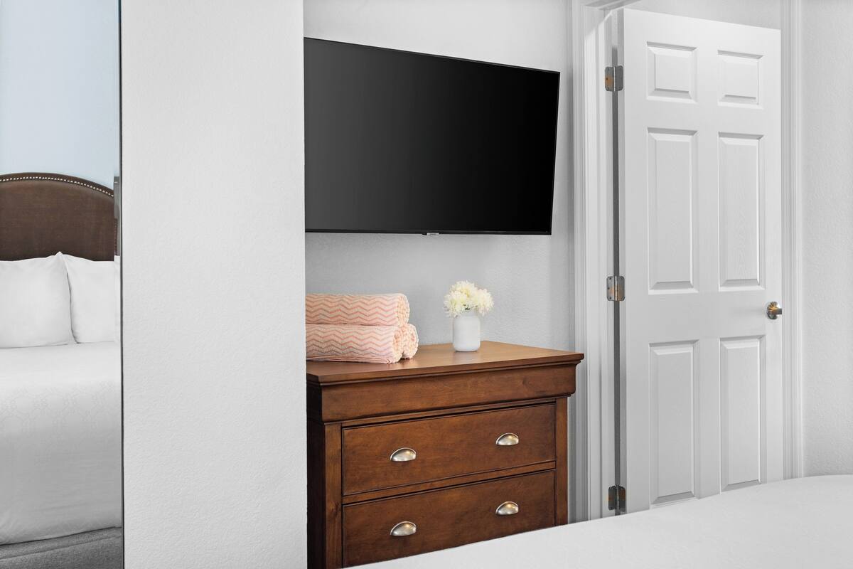 Ein Schlafzimmer mit einem Bett, einer Kommode mit Schubladen, einem flachbildfernseher, der über der Kommode angebracht ist, und einer weißen Tür.