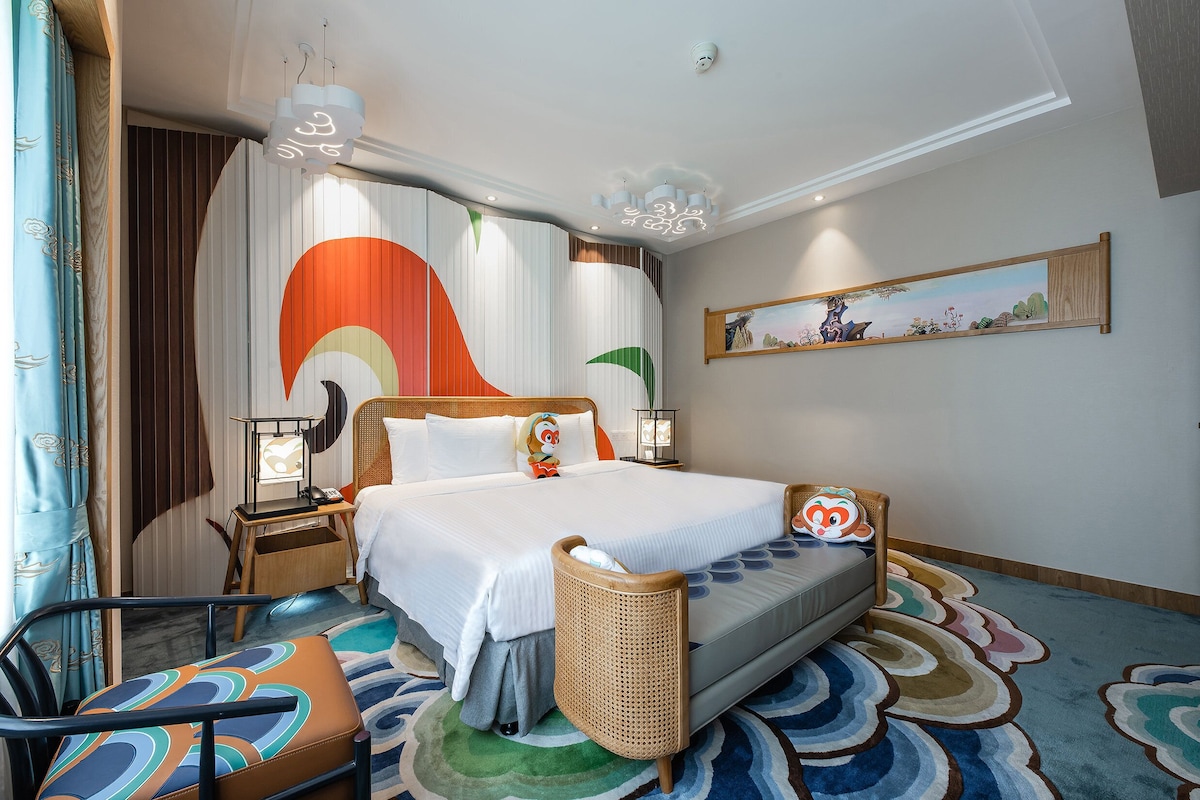 A hotel room with a large bed, a bedside table with a lamp, a chair, and a colorful rug.