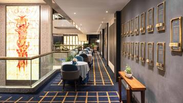 A hotel corridor with tables and chairs, a staircase, and a wall with plaques.