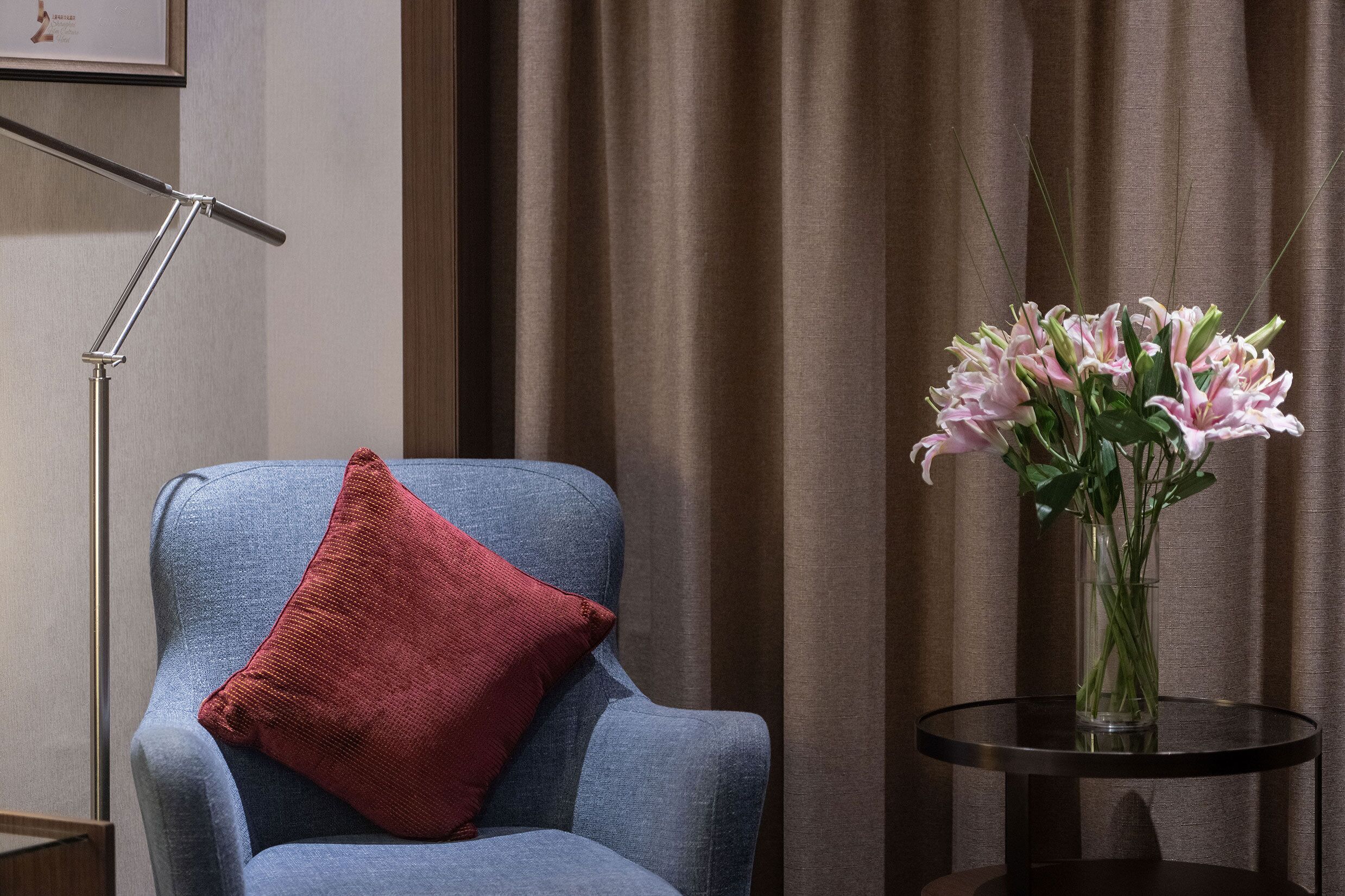 Une pièce avec un fauteuil bleu, un coussin rouge, une lampe sur pied et un vase rempli de fleurs roses, posé sur une petite table.