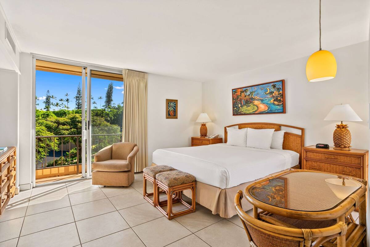 Ein Hotelzimmer mit Bett, Sessel, Tisch, einem Balkon mit Blick auf Bäume und Kunstwerken an den Wänden.