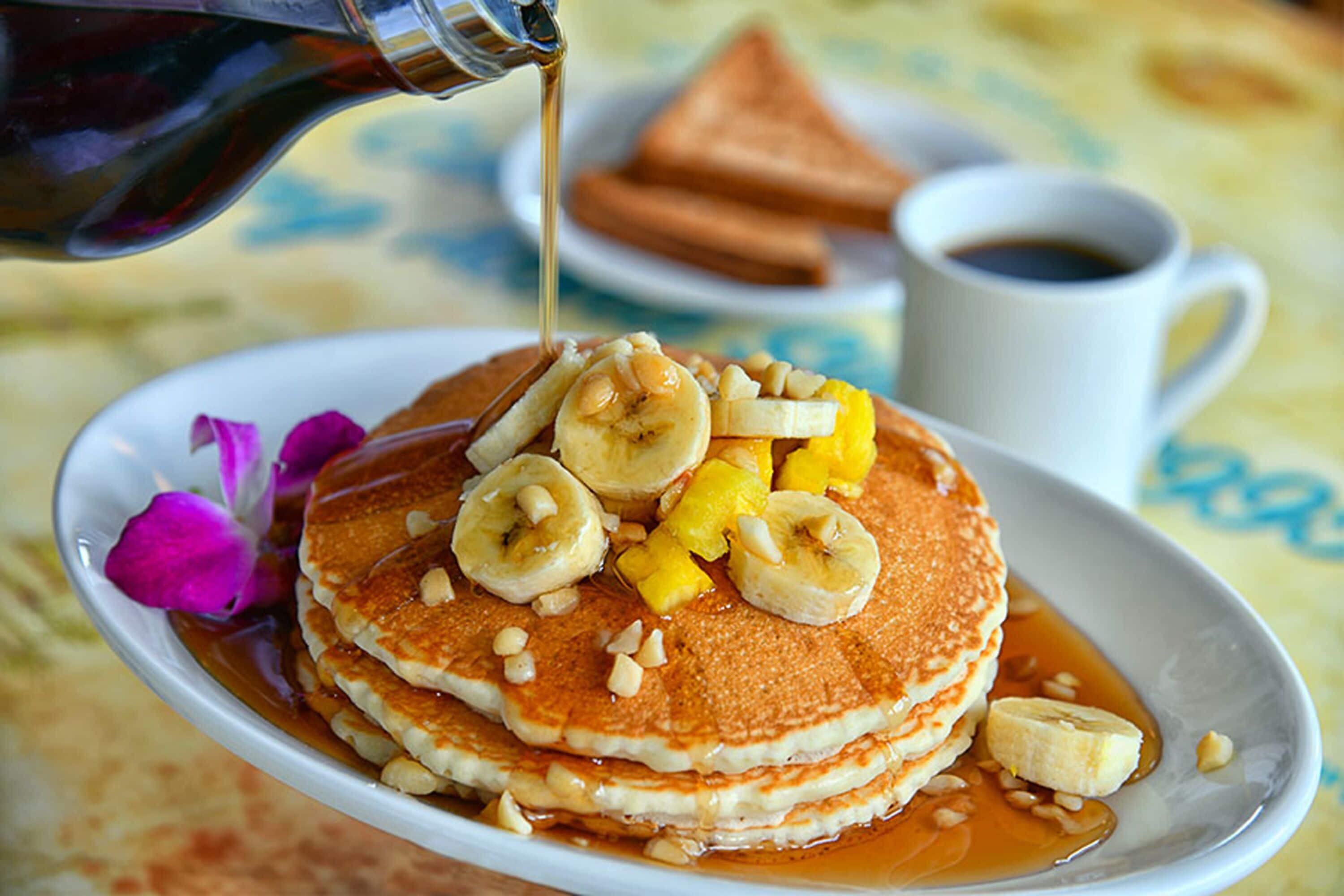 A plate of pancakes topped with bananas and syrup, with a cup of coffee and toast in the background.