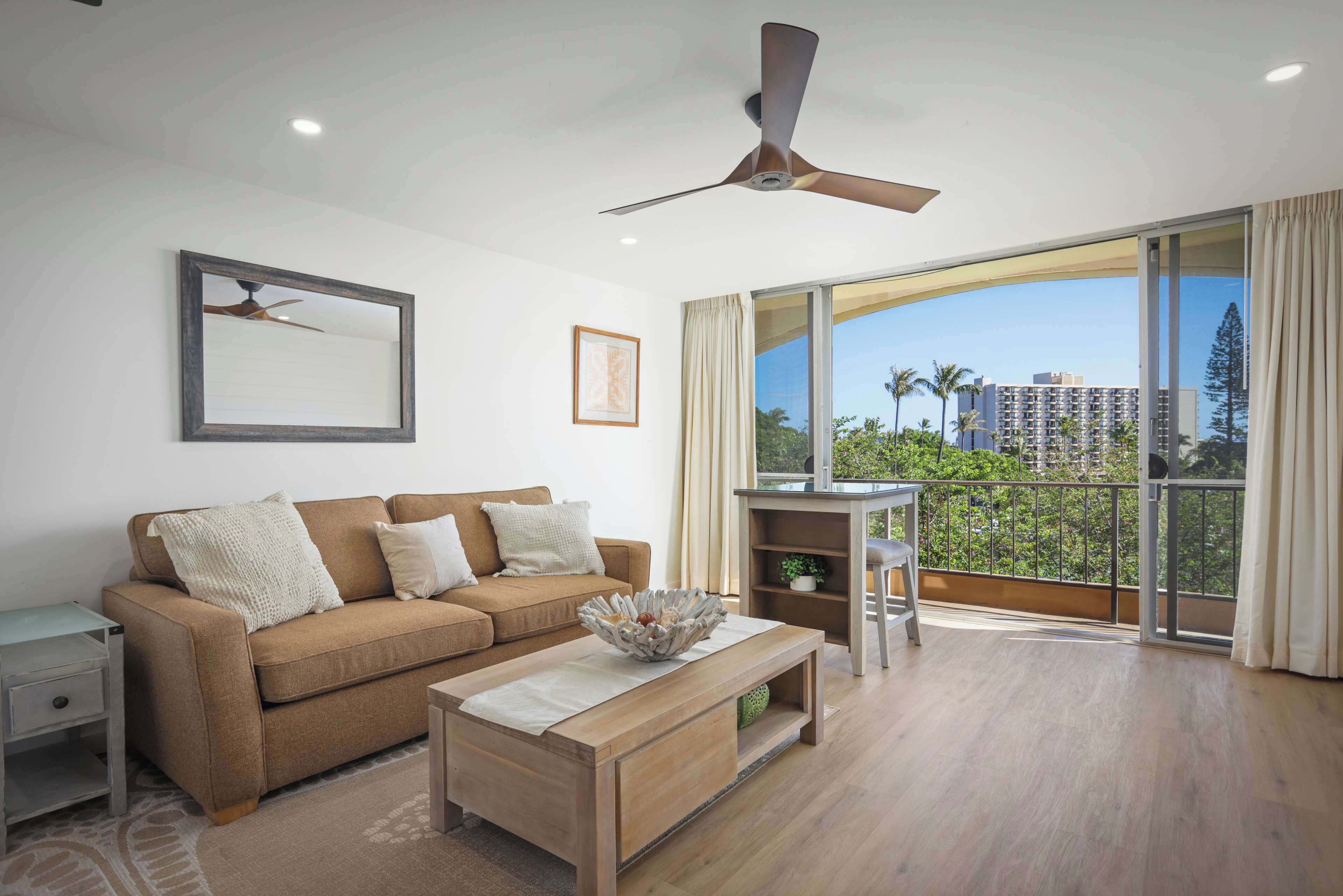 A modern living room with a sofa, coffee table, ceiling fan, and large windows offering a view of buildings and trees.