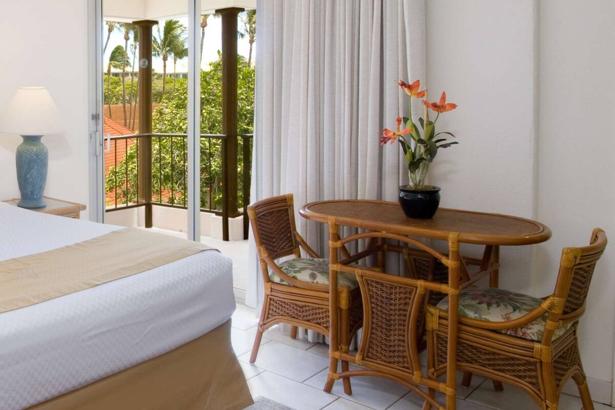 Ein Hotelzimmer mit Bett, Esstisch mit Stühlen, einer Zimmerpflanze und Blick auf Palmen vom Balkon aus.