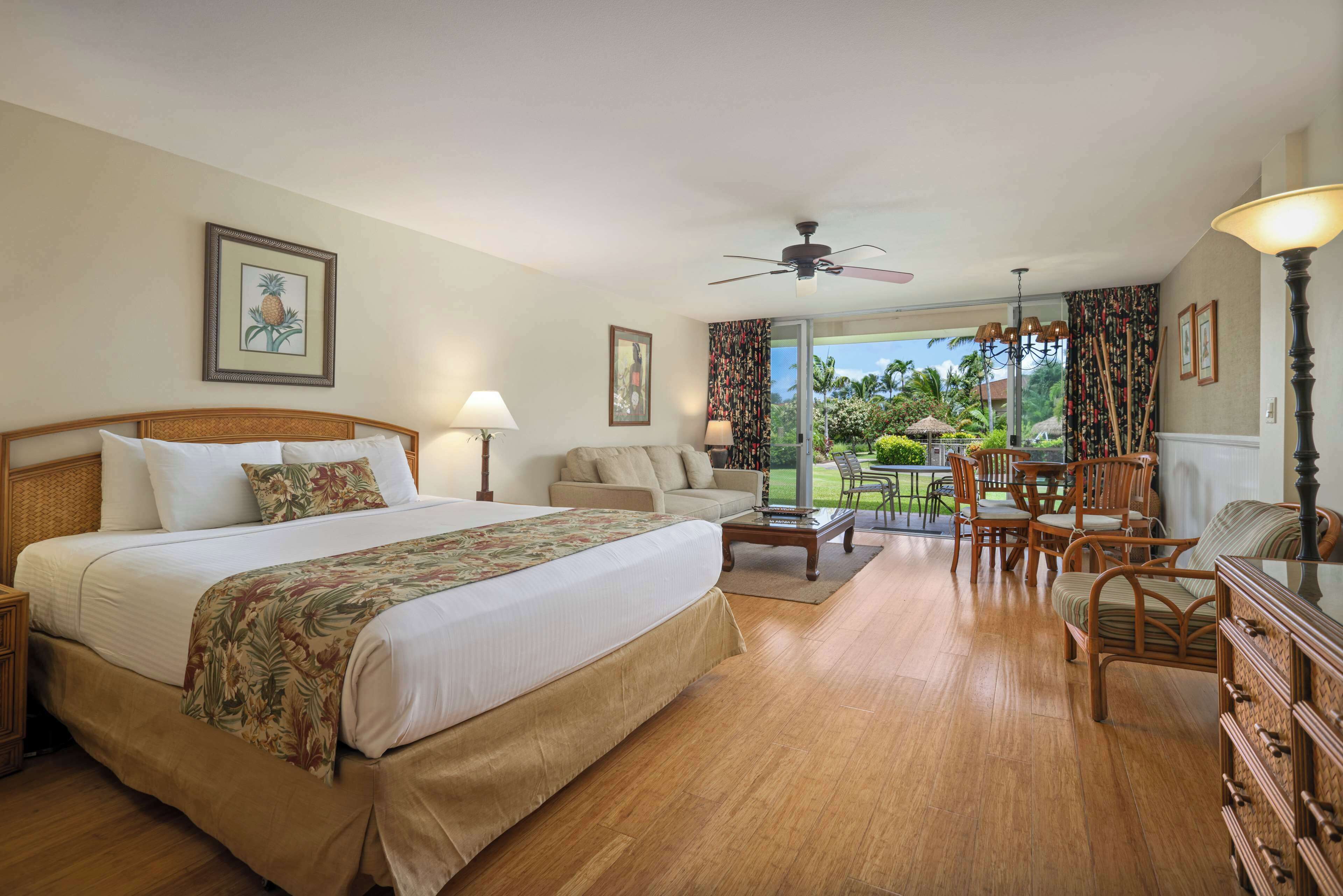 A hotel room with a bed, a sofa, a dining table with chairs, a ceiling fan, and a view of the outdoors.