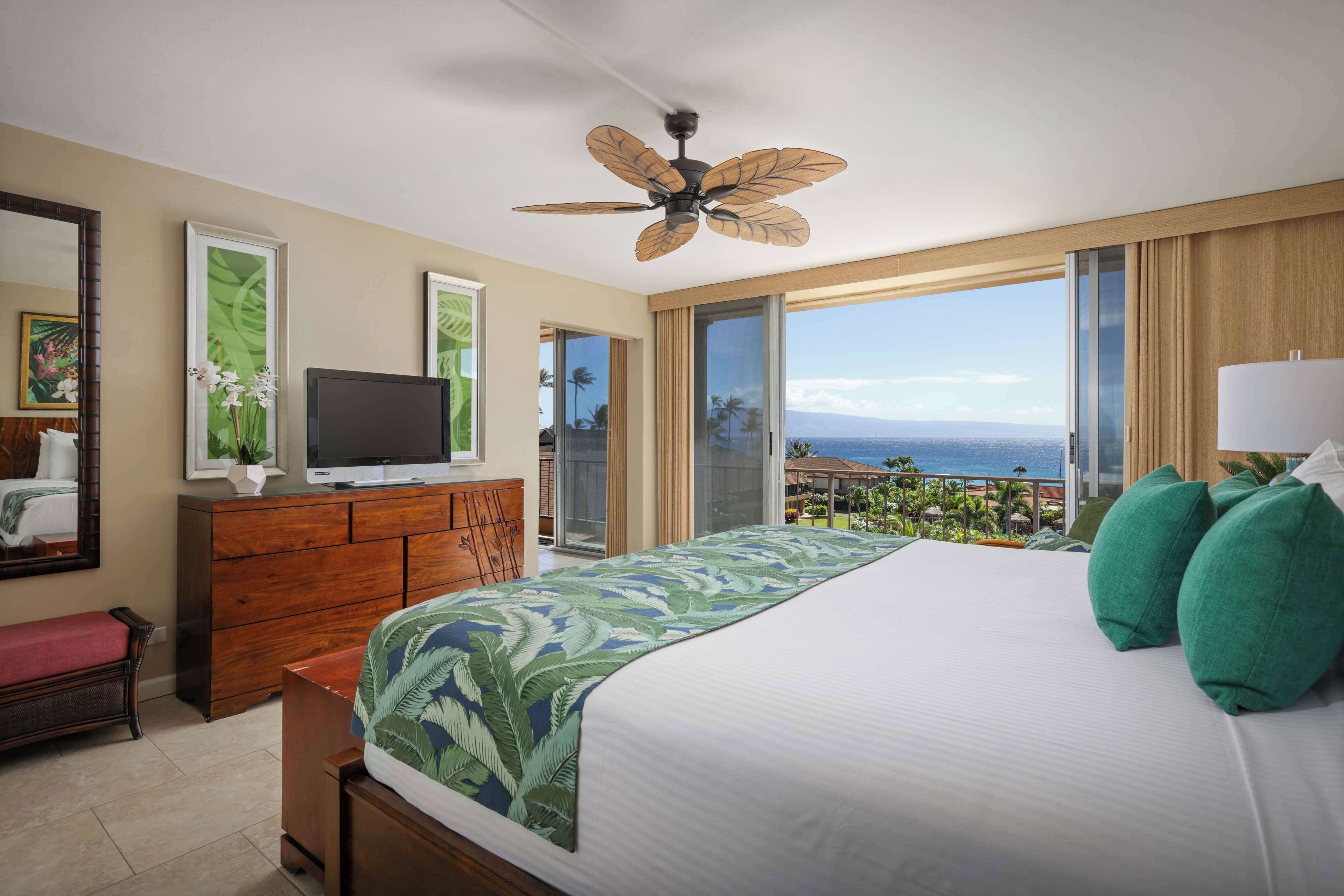 Habitación de hotel con cama, televisión, ventilador de techo, ventana grande con vistas y cómoda de madera.