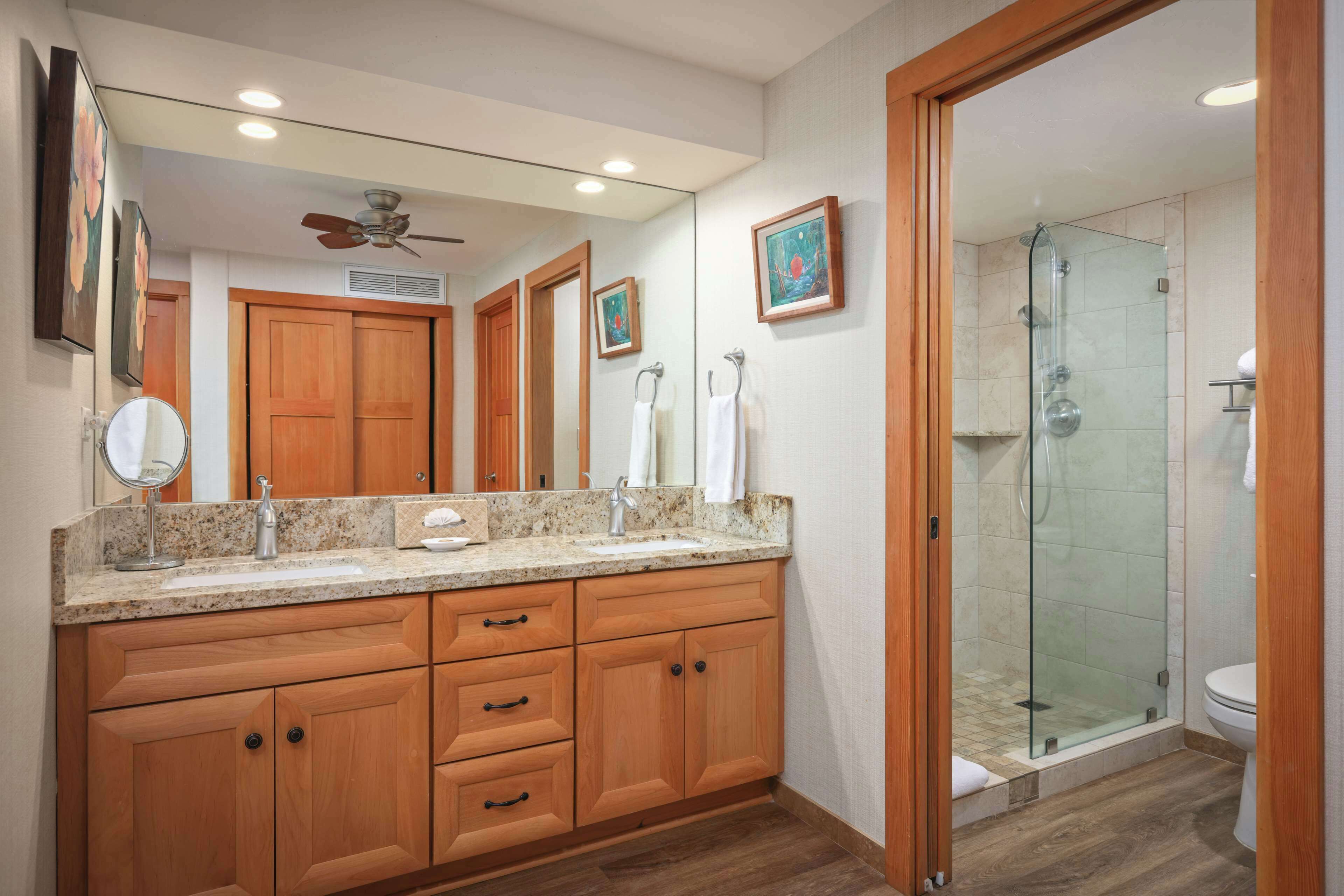 A bathroom with wooden cabinets, a large mirror, a ceiling fan, and a glass-enclosed shower.