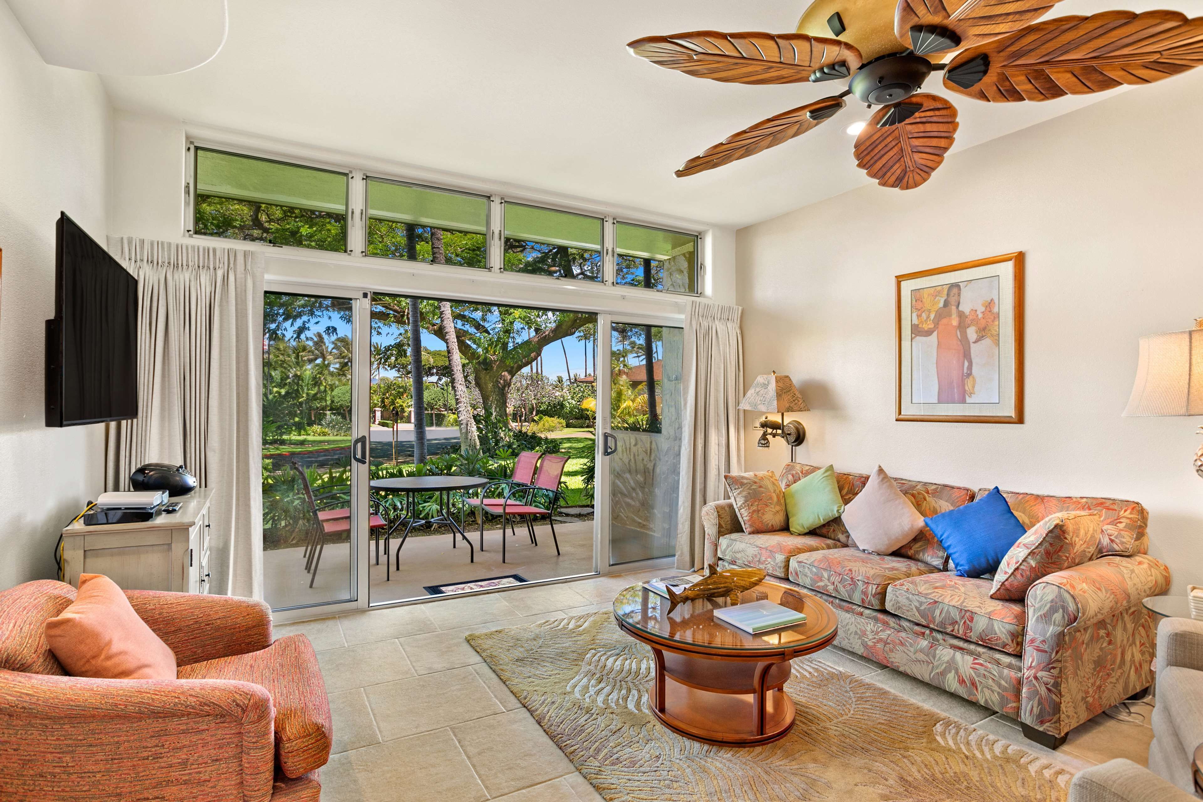 A living room with a sofa, coffee table, and a television. There is a large window with a view of greenery outside.