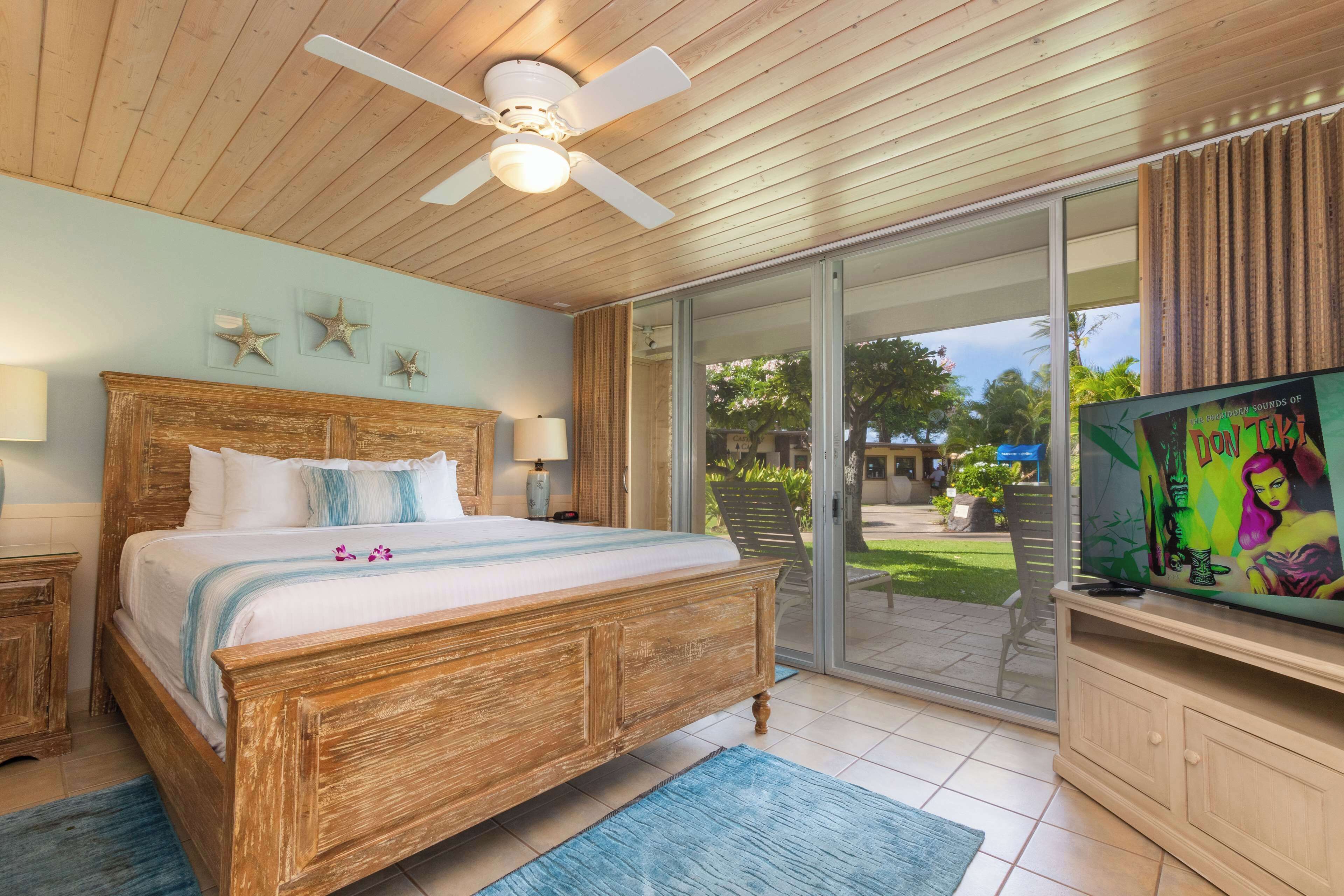 A bedroom with a wooden bed, a ceiling fan, a TV, and a sliding glass door to the outside.