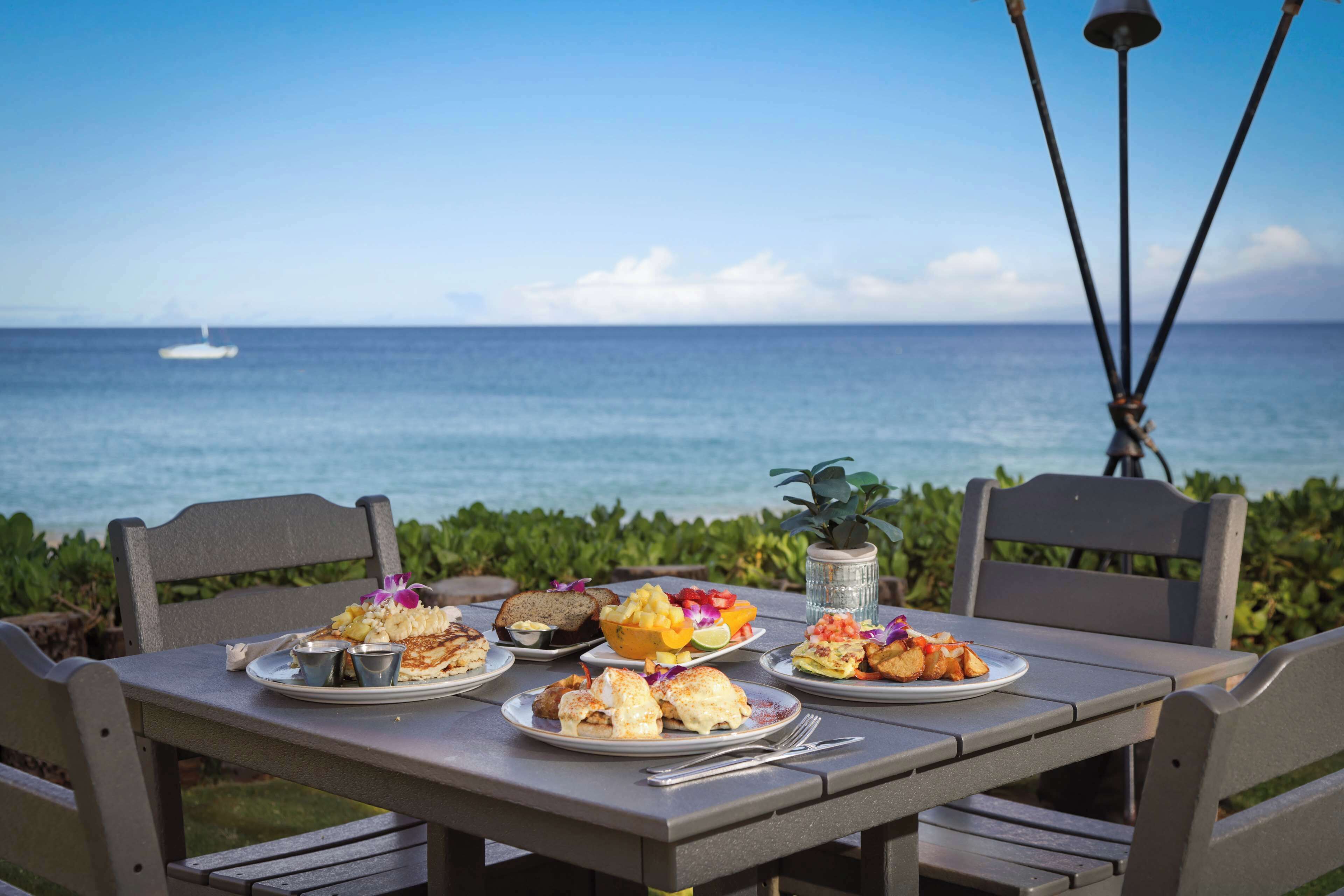Une table avec des assiettes de nourriture, une tasse en métal et une plante, le tout placé devant une vue sur l’océan.