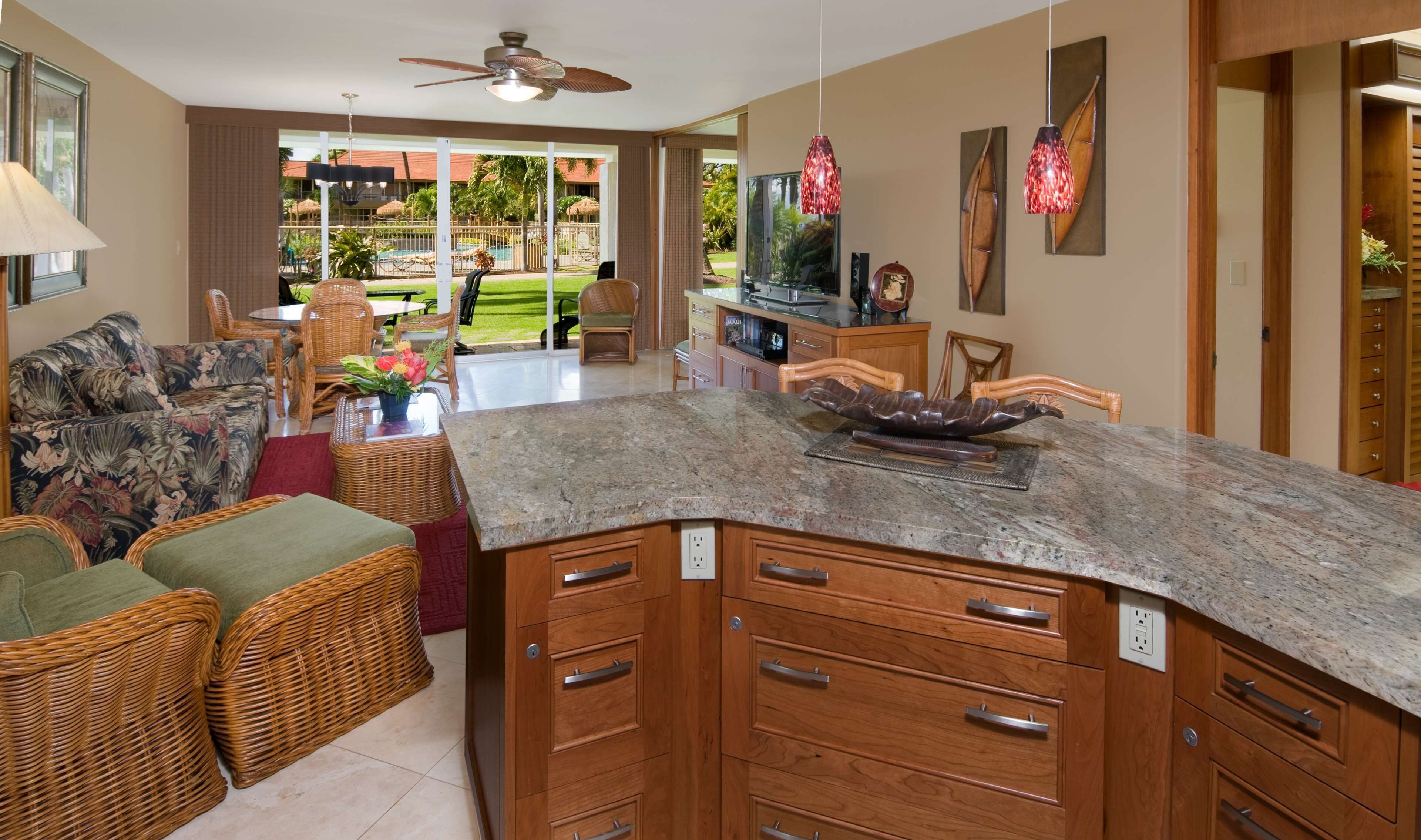 A kitchen with a granite countertop, wooden cabinets, wicker furniture, and a view of a pool and garden.