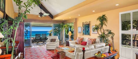 A spacious living area with a white sofa, red patterned rug, and a wooden coffee table. There are potted plants, a guitar, and a view of the ocean through large windows.