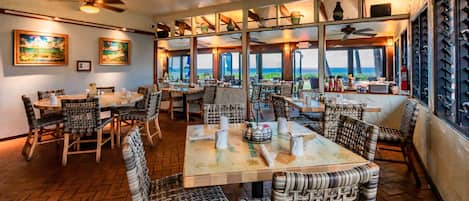 A restaurant with multiple tables and chairs, ceiling fans, and large windows offering a view.
