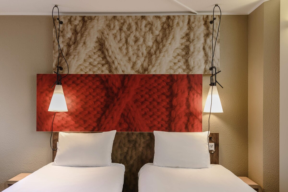 A hotel room with two beds, a red and brown textured wall, and two pendant lights.