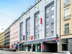 Un hotel de varios pisos con un llamativo letrero rojo de 'ibis', una entrada visible y un cartel de estacionamiento que indica un límite de velocidad de 34 km/h.