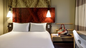 Une chambre d’hôtel avec un grand lit, deux lampes de chevet, une table de chevet en bois, une chaise et une tête de lit fixée au mur.