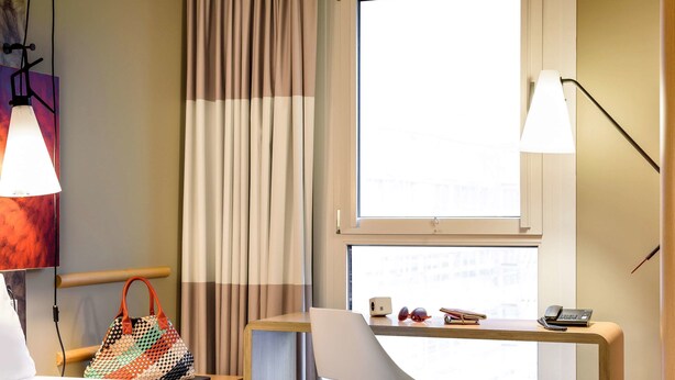 Una camera d'albergo con un letto, una scrivania con telefono e occhiali da sole, una borsa e una finestra con tende.